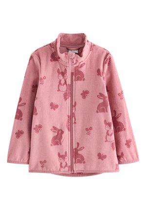 Fille rousse souriante portant une veste polaire rose à motifs animaux, un jean bleu, des baskets colorées et des cache-oreilles blancs et duveteux, debout sur fond blanc.