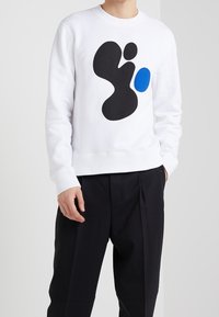 Sweat-shirt blanc avec un motif abstrait noir et un accent bleu. Le tissu semble doux et présente une finition texturée.