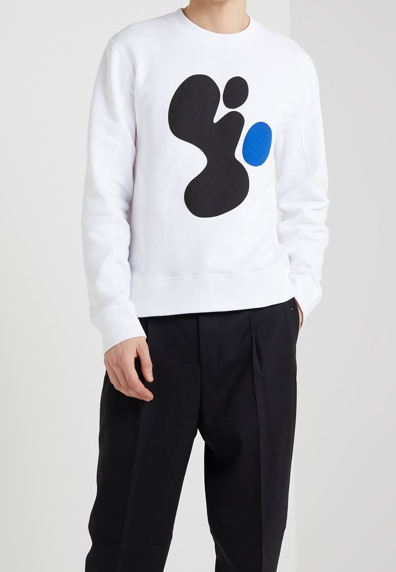 Sweat-shirt blanc avec un motif abstrait noir et un accent bleu. Le tissu semble doux et présente une finition texturée.