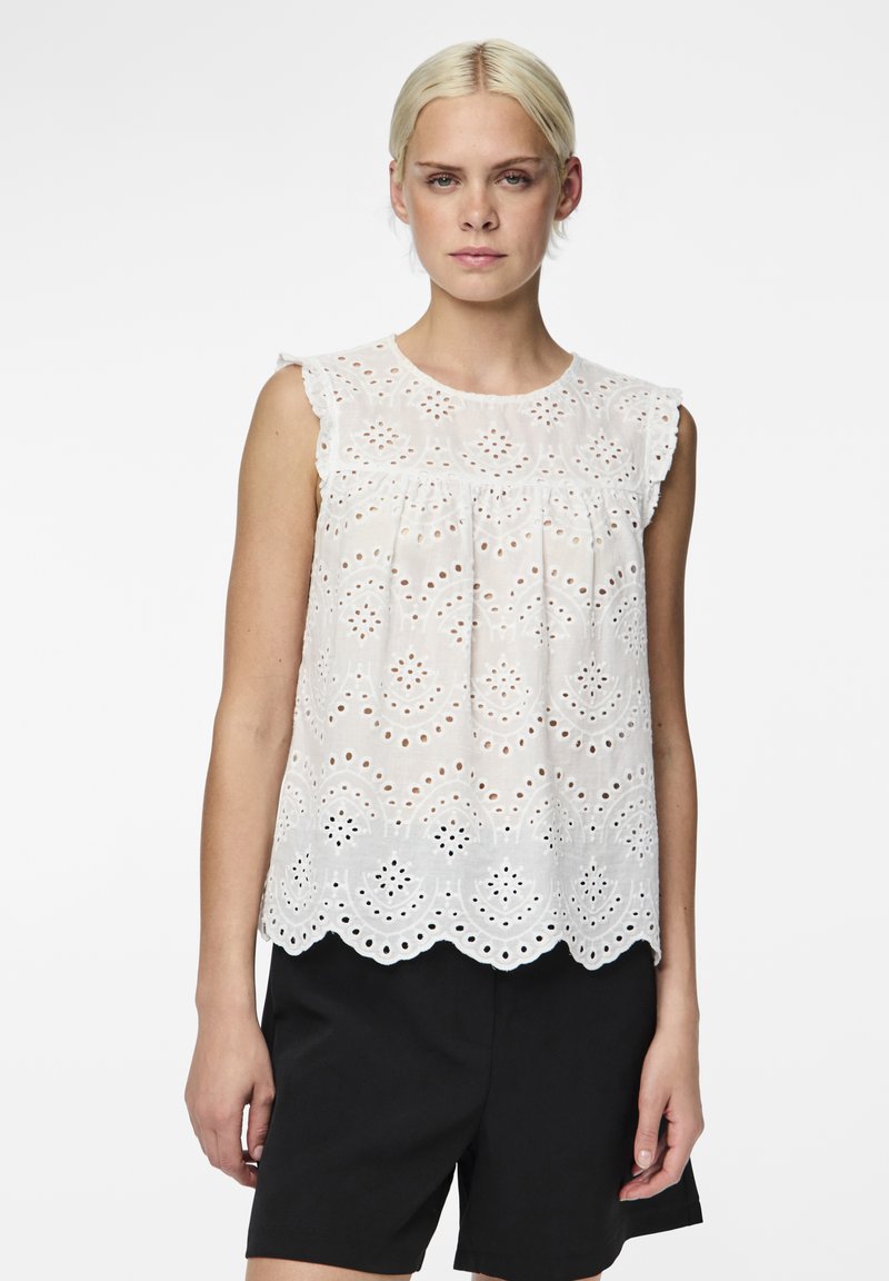 Haut sans manches blanc avec des motifs découpés complexes, des emmanchures froncées et un ourlet festonné, associé à un short noir.