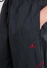 Melnas šortas, izgatavoti no viegla, krokota auduma, ar elastīgu jostasvietu un sarkanu izšūtu logo uz kreisās kājas.