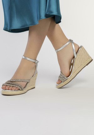 Sandalias de cuña plateadas con tiras decorativas metálicas, tacón de yute trenzado y correa en el tobillo. Diseño de punta abierta con un acabado texturizado.