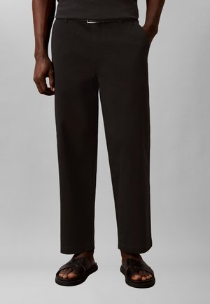 Muž nosící černé kalhoty s přímými nohavicemi a bočními kapsami, černý pásek a černé kožené sandály na jednobarevném šedém pozadí.