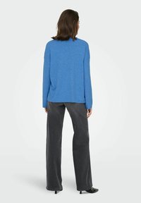 Pull en tricot bleu à manches longues, coupe décontractée, col rond, associé à un pantalon large gris foncé et des chaussures noires.
