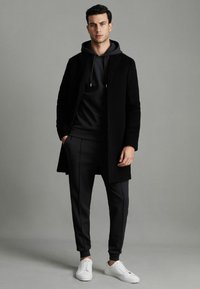 Palton negru din lână peste un hanorac negru și pantaloni jogger asortați. Accesorizat cu adidași albi. Îmbrăcămintea are o siluetă modernă, elegantă.