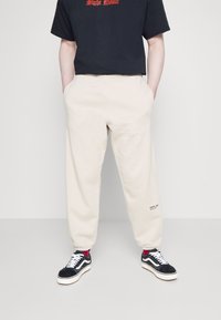 Pantalones de chándal beiges con puños elásticos y bolsillos laterales, con un pequeño logo "REPLAY" en la pierna izquierda, combinados con zapatillas negras.