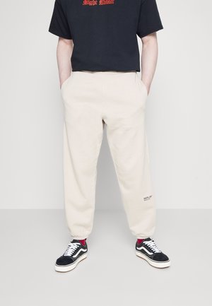 Pantaloni della tuta beige con polsini elastici e tasche laterali, con un piccolo logo "REPLAY" sulla gamba sinistra, abbinati a sneaker nere.