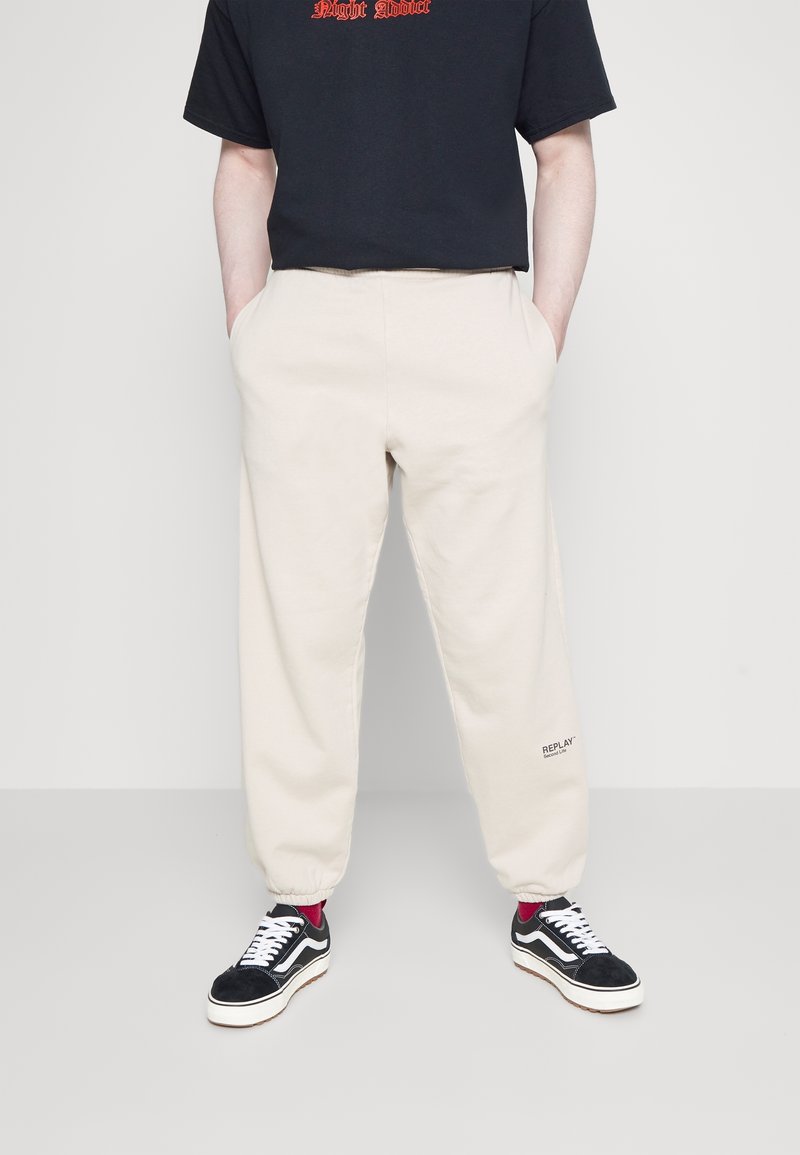 Pantalones de chándal beiges con puños elásticos y bolsillos laterales, con un pequeño logo "REPLAY" en la pierna izquierda, combinados con zapatillas negras.