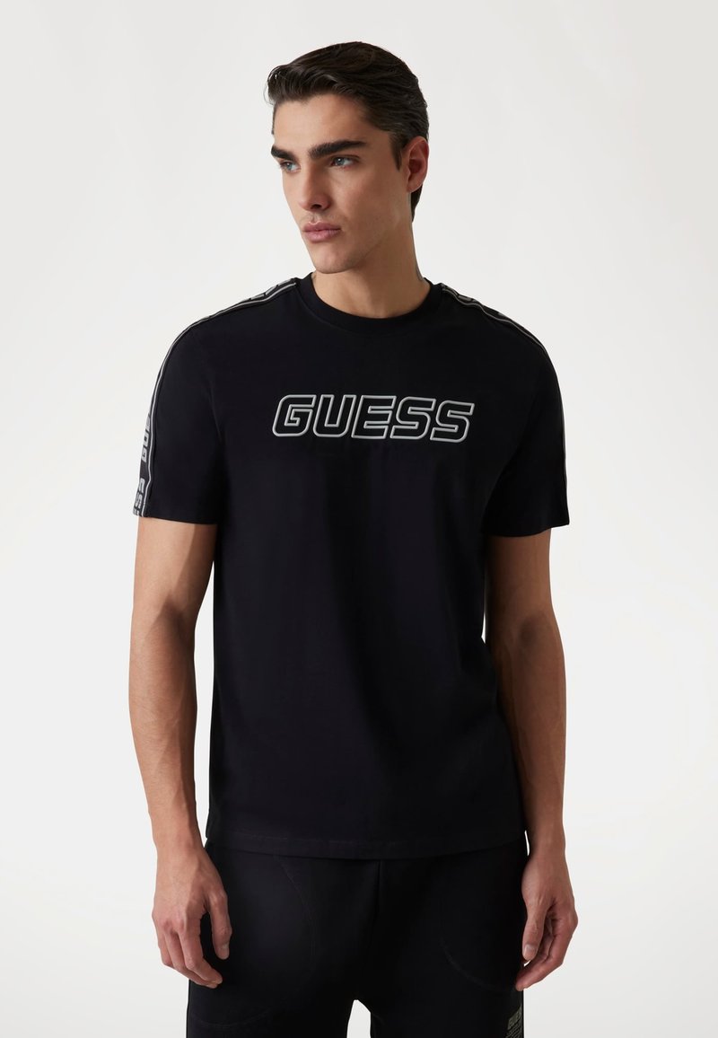 Schwarzes Baumwoll-T-Shirt mit einem großen weißen "GUESS"-Logo auf der Vorderseite und kontrastierenden Seitestreifen. Kurzärmeliges Design mit Rundhalsausschnitt.