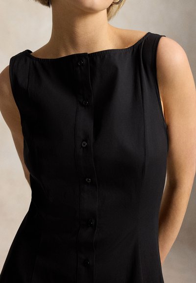 Kvinne iført en ermeløs svart kjole med knapper og bred halslinning, samt store øredobber, stående mot en nøytral bakgrunn.