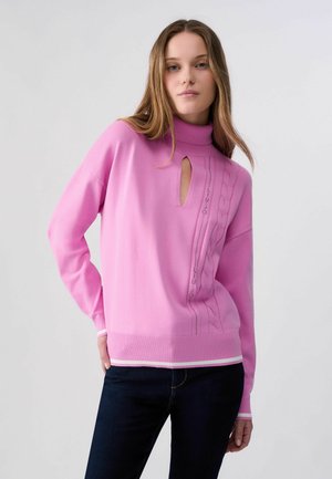 Mujer de pie de frente, vistiendo un jersey de cuello alto rosa de manga larga con abertura tipo ojo de cerradura y vaqueros oscuros, sobre un fondo liso.
