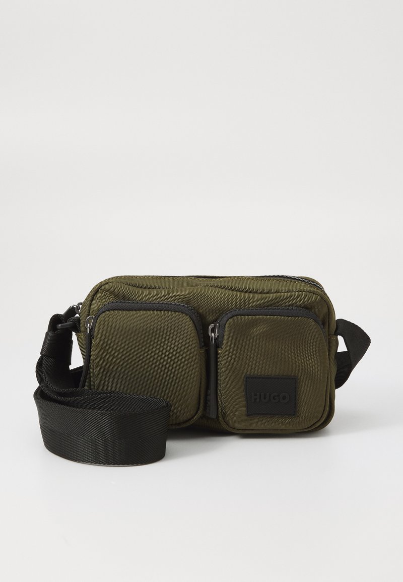 Olivgrüne Stoff-Crossbody-Tasche mit zwei vorderen Reißverschlusstaschen, verstellbarem schwarzen Tragegurt und schwarzem Logo-Patch. Rechteckige Form mit strukturierten Oberflächen.