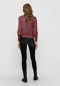 Donna con capelli ondulati che indossa un maglione a maniche lunghe mauve, jeans neri aderenti e stivali neri alla caviglia, in piedi davanti a uno sfondo semplice.