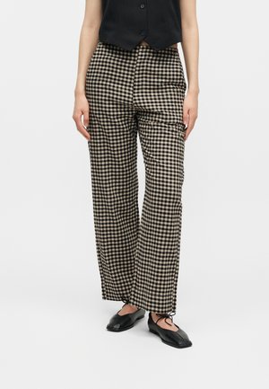 Osoba v čierno-béžových kockovaných nohaviciach s vysokým pásom a širokými nohavicami, čiernej zapínacej veste a čiernych šnurovacích balerínach na bielom pozadí.