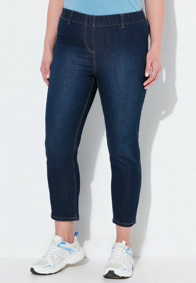 Tummansiniset farkut, joissa on joustava vyötärö, slim fit -malli ja nilkkapituiset lahkeet. Viimeistelytikkaus näkyy saumoissa.