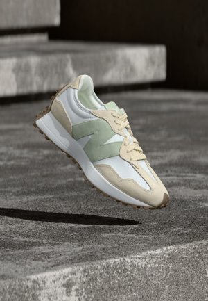 Baskets beige et vert clair avec semelle blanche et lacets, flottant au-dessus d'une surface en béton texturée avec des marches en arrière-plan.