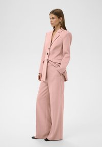 Rosa maßgeschneiderter Anzug mit langen, weiten Hosen und einengendem Blazer. Verfügt über knopfartige Doppelknöpfe und ein elegantes Design. Glatter, strukturierter Stoff.