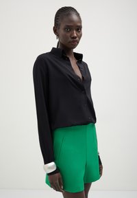 Maglietta nera a maniche lunghe con chiusura a bottoni, abbinata a pantaloni corti alti di un vivace verde. Presenta un tessuto elegante e un accento bianco a contrasto sui polsini.