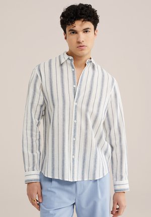 Mladý muž oblečený v bielej košeli s dlhými rukávmi, zapínanej na gombíky, s tenkými vertikálnymi sivými pruhmi a svetlomodrých nohaviciach, stojaci pred jednofarebným pozadím.