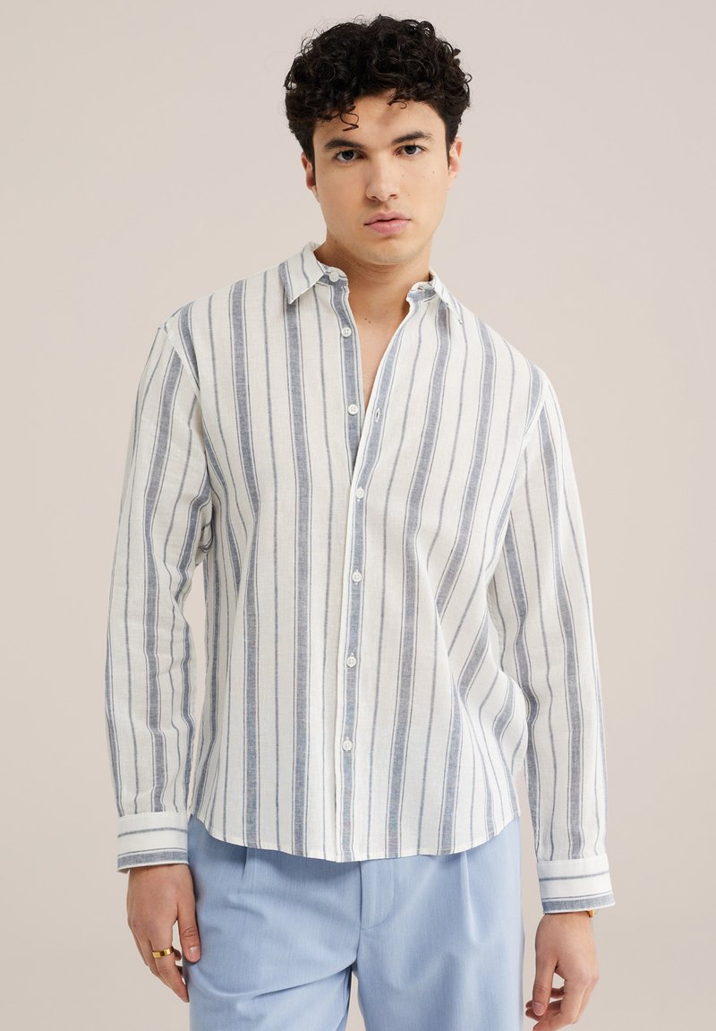 Jeune homme portant une chemise blanche à manches longues avec de fines rayures verticales grises et un pantalon bleu clair, debout devant un fond uni.
