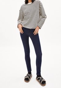 Gestreiftes Langarmshirt in Schwarz und Weiß, kombiniert mit dunkelblauen Skinny Jeans und schwarzen Sandalen mit hellen Akzenten und einer Holzsohle.