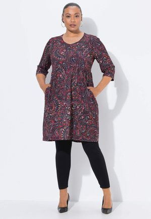 Frau steht mit Händen in den Taschen, trägt ein mehrfarbiges Paisleymuster-Kleid, schwarze Leggings, schwarze Absätze und Ohrhänger vor einem weißen Hintergrund.