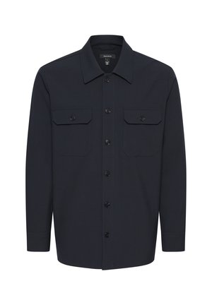 Mężczyzna noszący granatową kurtkę zapinaną na guziki z patkami, dopasowane wąskie spodnie, czarne mokasyny i białą koszulkę na jednolitym tle.