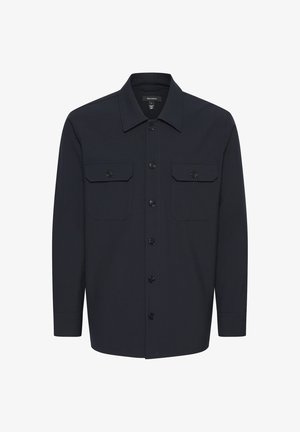 Mand iført navy knappet jakke med klaplommer, matchende slim-fit bukser, sorte loafers og en hvid T-shirt mod en ensfarvet baggrund.