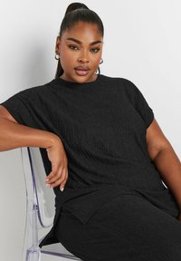 Czarna, fakturowana bluzka o luźnym kroju, z krótkim rękawem i zaokrąglonym dołem. Materiał ma marszczony wzór, co dodaje atrakcyjności wizualnej.