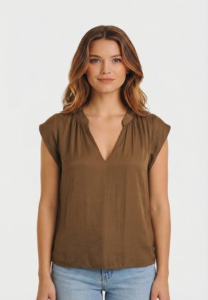 Mujer con cabello castaño ondulado, que lleva una blusa de manga corta verde oliva con cuello en V y vaqueros azul claro, de pie frente a un fondo blanco liso.