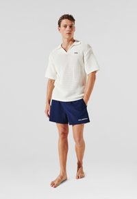 Polo alb texturat cu mâneci scurte, asortat cu pantaloni scurți bleumarin cu logo alb. Modelul este desculț, stând pe un fundal simplu.