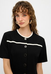 Vestido corto negro de manga corta con cuello redondo, franjas horizontales blancas y botones en un tono dorado en contraste. Textura suave, ajuste estructurado.