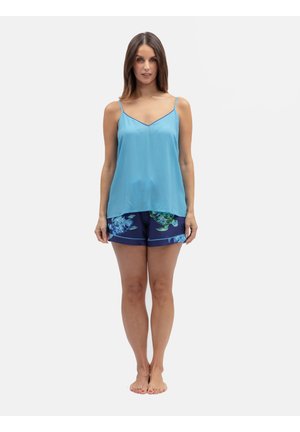 Vrouw staat op blote voeten, draagt een lichtblauwe mouwloze top en marineblauwe korte broek met groene en blauwe schildpaddenprints.