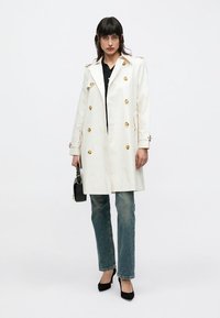 White trench coat with gold buttons, double-breasted design, shoulder epaulets, and a straight cut. Worn over black top and denim.