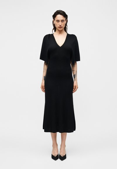Femme portant une robe midi noire à col en V et manches courtes style cape, debout droite devant un fond clair uni.