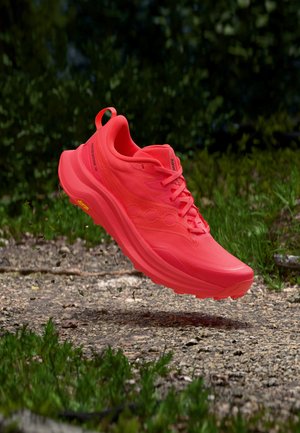 Chaussure de trail running rouge vif en suspension au-dessus d'un chemin de gravier avec de l'herbe verte et du feuillage en arrière-plan.