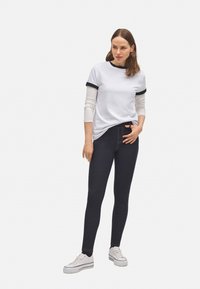 Camiseta blanca con ribete negro, superpuesta sobre una camisa de manga larga. Jeans oscuros ajustados con costuras naranjas y zapatillas blancas.