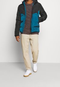 Gesteppten Jacke aus schwarzem und türkisfarbenem Stoff, mit Reißverschlusstaschen und Kapuze. Getragen über einem strukturierten braunen Pullover und beigen Hosen, mit weißen Turnschuhen.