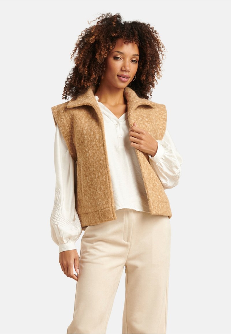 Tan wool blend sleeveless vest with a collar, textured surface, worn over a white button-up shirt, paired with light beige trousers.