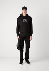Black sweatshirt with graphic logo, paired with black pants and black sneakers. Holding headphones, wearing a black cap. Simple design.