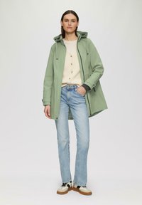 Chaqueta impermeable verde claro con capucha, puños y botones negros, llevada sobre un suéter de punto color crema, combinada con jeans azul claro y zapatillas beige.