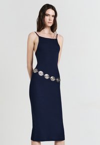 Femme aux longs cheveux foncés portant une robe midi sans manches bleu marine avec des fines bretelles et une ceinture décorative en argent.