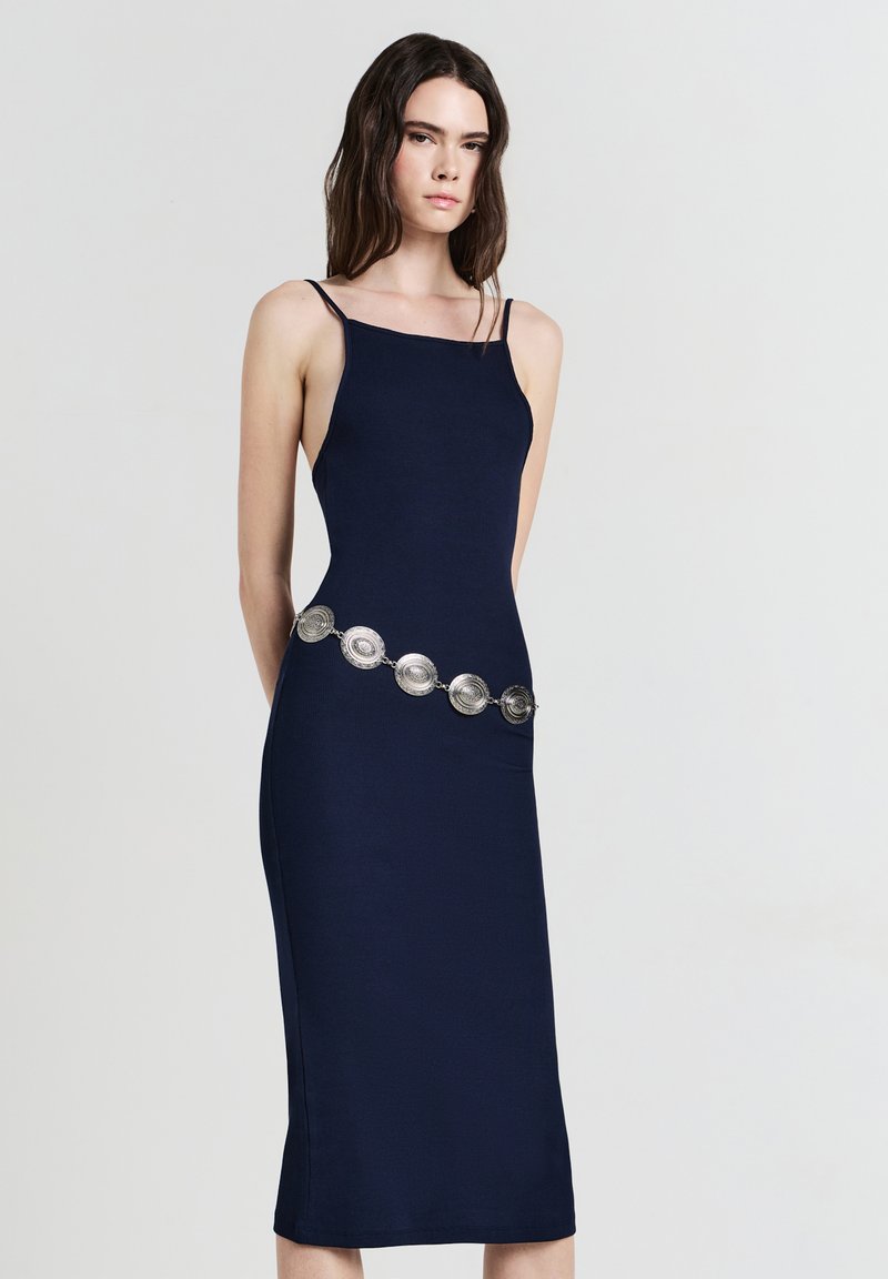 Femme aux longs cheveux foncés portant une robe midi sans manches bleu marine avec des fines bretelles et une ceinture décorative en argent.