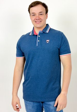 Jeune homme souriant portant un polo bleu avec un col et des bords de manches rayés, et un jean bleu clair, debout devant un fond blanc.