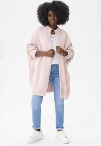 Jasnoróżowy, oversize sweter z wysokim kołnierzem, ściągaczami oraz dwiema kieszeniami, noszony na białej bluzce i zestawiony z jasnoniebieskimi dżinsami.