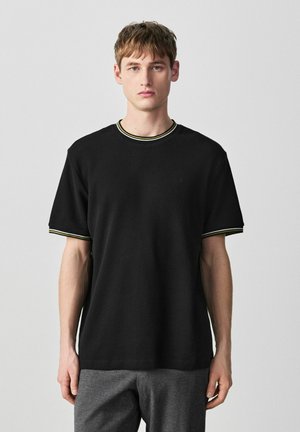 Fiatal férfi fekete rövid ujjú ingben, melynek gallérján és ujjain fehér és sárga csíkos díszítés található, egy szürke, egyszínű háttér előtt áll.