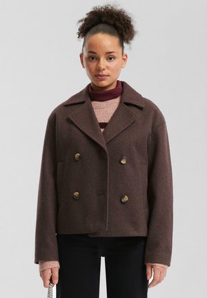 Veste en mélange de laine marron à double boutonnage avec de grands boutons, revers en pointe et poches avant. Portée par-dessus un pull col roulé bordeaux.
