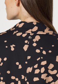 Vêtement noir avec des formes abstraites beiges, présentant un col et un bouton décoratif à l'arrière. Texture lisse, design ajusté.