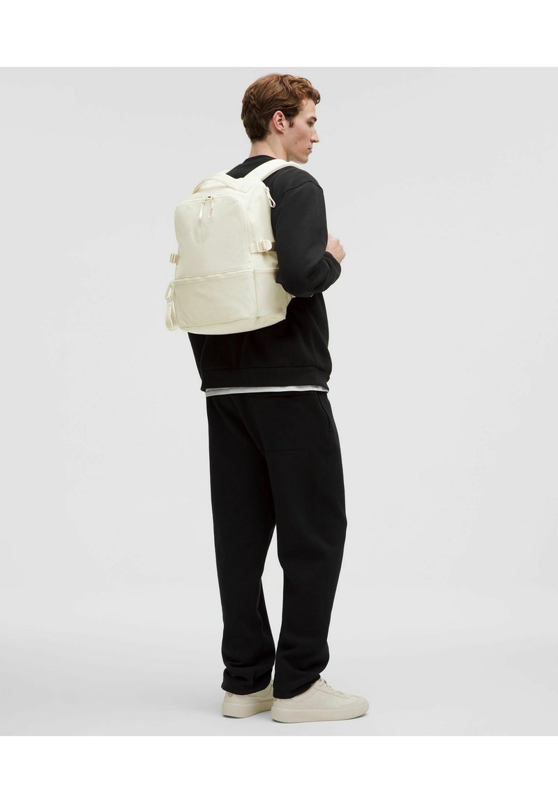Mochila blanca con doble tirantes, bolsillo frontal y hebillas laterales ajustables, llevada sobre un atuendo negro. Textura suave, diseño minimalista.