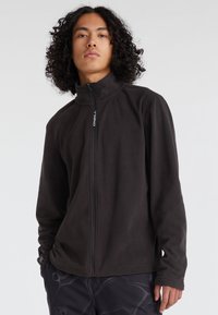 Schwarze Fleecejacke mit hohem Kragen und voller Reißverschluss. Besitzt ein Logo auf der Vorderseite und strukturierter Stoff. Das Model trägt dunkle Hosen.